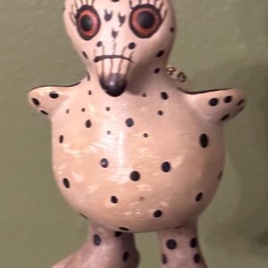 May include: A whimsical, hand-painted ceramic figurine of a bird-like creature. The figure is beige with black spots, red eyes, and a dark beak. It has small arms and large feet, and is suspended by a green string.