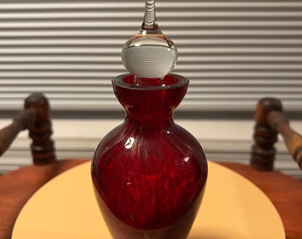 May include: A red glass bottle with a clear glass stopper. The bottle has a textured surface and a rounded bottom. The stopper has a long, thin neck and a round top.