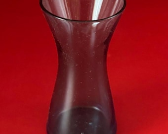 May include: A clear, dark-tinted glass vase with a flared top and a gently curved body. The vase is empty and sits against a solid red background. The glass appears to be smooth and transparent.