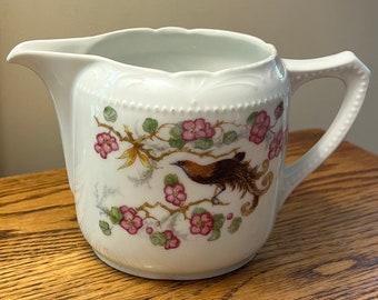 May include: White porcelain creamer with a floral design featuring pink flowers and a brown bird with a long tail.
