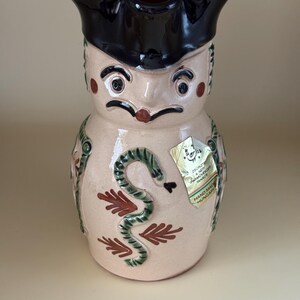 May include: A ceramic pitcher with a face design, featuring a black brim, eyes, and a mustache. The pitcher has a snake and leaf design, with a "Hand Made Hungary" tag.