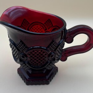 May include: A red glass creamer with a decorative pattern and a handle. The creamer has a footed base.