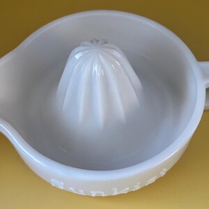 May include: A white ceramic citrus juicer with a built-in reamer and a pouring spout. The juicer has a handle and is set against a yellow background. The reamer is cone-shaped with ridges.