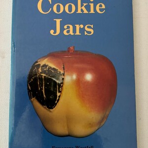 Peut inclure: Un livre intitulé "An Illustrated Value Guide To Cookie Jars" avec un pot à biscuits en forme de pomme rouge et jaune sur la couverture. Le livre est écrit par Ermagene Westfall.