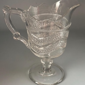 May include: Clear glass pitcher with a decorative handle and a scalloped rim. The pitcher features a detailed pattern of diamond shapes and horizontal bands. It stands on a round base and is suitable for serving beverages.