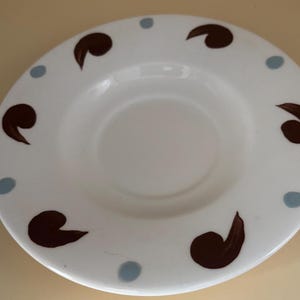 Op de afbeelding: Witte keramische schotel met een bruin en blauw ontwerp. De schotel heeft een ring van bruine kommavormen rond de rand en blauwe stippen tussen elke komma.