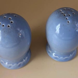 May include: A pair of light blue ceramic salt and pepper shakers. Each shaker has a rounded top with several holes and sits on a decorative base. The shakers have a glossy finish and a subtle color gradient.