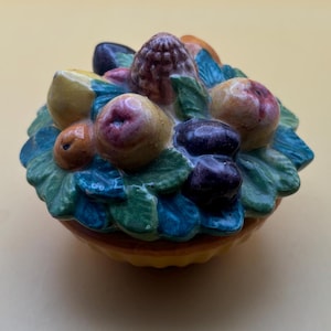 May include: A ceramic fruit basket with a yellow base and green leaves. The basket is filled with colorful fruit, including red apples, yellow lemons, and purple grapes.