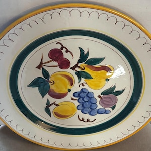 May include: Oval ceramic serving platter with a hand-painted fruit design. The platter features a yellow rim, a green border, and a white center with illustrations of cherries, pears, grapes, and plums. The edge has a scalloped design.