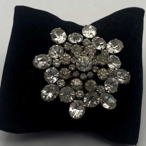 May include: A silver-toned brooch with a cluster of clear, faceted rhinestones. The brooch is arranged in a floral or starburst pattern, with various sizes of stones. The brooch is displayed on a black velvet surface.