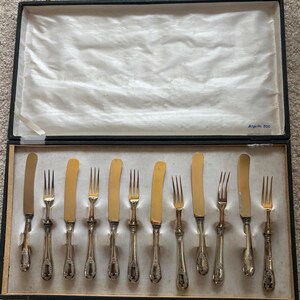 May include: A set of antique silverware, including butter knives and forks, displayed in a black box with a white interior. The knives have gold-colored blades and ornate silver handles. The box is labeled "Argento 800".