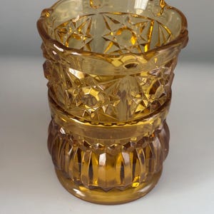 May include: Amber-colored glass vase with a detailed, textured design. The vase features a scalloped rim and geometric patterns, including starburst and diamond shapes. The base has vertical ridges, adding to its vintage aesthetic.