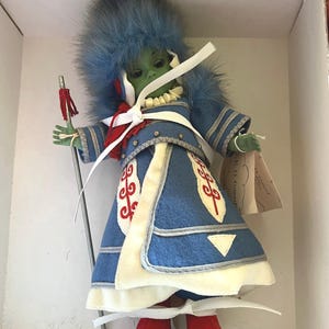May include: A green-skinned doll wearing a blue and white outfit with red accents. The doll is wearing a blue fur hat and holding a staff with red tassels. The doll is standing on a white base.