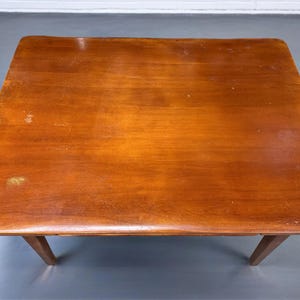 May include: A square wooden table with a warm, medium-brown finish and visible wood grain. The tabletop features a smooth surface with minor signs of wear and light scuffing. It is supported by four tapered legs extending from the corners.