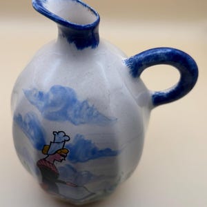 May include: A white ceramic pitcher with a blue handle and spout. The pitcher has a blue and white painted design of a woman in a blue and white dress and hat. The pitcher is approximately 15 cm tall.