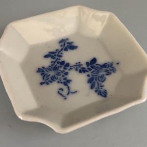 May include: A small, square, white ceramic dish with a blue floral design. The dish has a slightly scalloped edge and is likely used for serving small portions or as a decorative item. The blue floral pattern adds a touch of elegance.