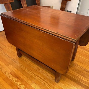May include: A dark brown wooden drop leaf table with a smooth, polished surface. The table has a rectangular top with two drop leaves that can be extended to create a larger surface area. The table is supported by four legs.