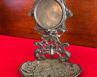 Puede incluir: Un marco de fotos de metal plateado con un diseño decorativo. El marco tiene una abertura redonda para una foto y una base con un diseño decorativo. El marco está sobre una superficie roja.