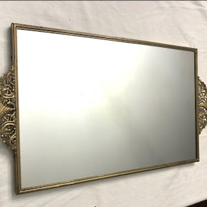 May include: A gold-toned rectangular tray with a mirrored surface and ornate, decorative edges. The tray has a vintage style.