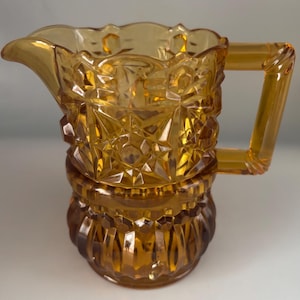 May include: Amber-colored glass pitcher with a geometric pattern. The pitcher has a spout, a handle, and a decorative design. The pitcher is approximately 6 inches tall.