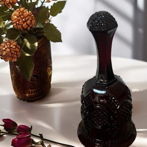 Puede incluir: Una campana de cristal burdeos oscuro con intrincados patrones en relieve. La campana tiene una base redondeada y un cuello largo y delgado, rematado con un asa esférica texturizada. Junto a la campana hay un jarrón con flores decorativas y unas rosas rojas.