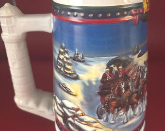Könnte beinhalten: Ein weißer Bierkrug aus Keramik mit Henkel. Der Krug zeigt eine Winterszene mit einer Pferdekutsche und einer verschneiten Landschaft. Der Griff ist weiß und hat ein dekoratives Design. Der obere Rand ist weiß, und die Basis hat einen rot-blauen Rand.