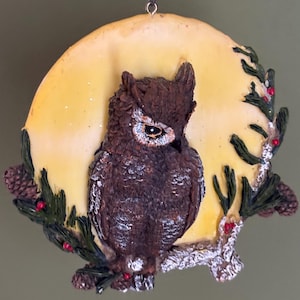 May include: A brown and white owl ornament with a yellow moon background. The owl is perched on a branch with green pine needles and red berries.