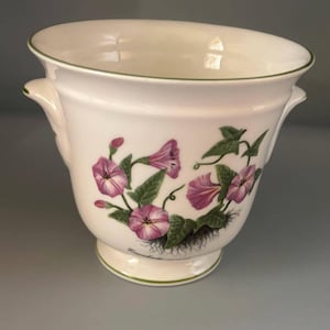 May include: White ceramic planter with a green rim and handles. The planter features a floral design of pink morning glories and green leaves. The base of the planter has a small foot and a green trim.