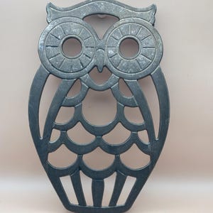 May include: A black metal owl-shaped hot pad with a cut-out design. The owl has large eyes and a detailed body.
