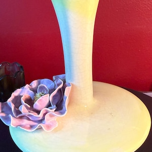 May include: A pale yellow ceramic vase with a wide, flat base and a slender, flared neck. A large, lavender and pink ceramic flower is attached to the vase. The vase has a crackle glaze finish.