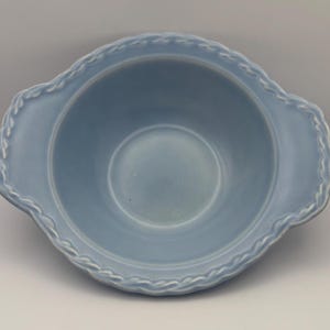 May include: A light blue ceramic bowl with a scalloped rim and two small handles. The bowl has a smooth, glossy finish and a circular interior. The rim features a decorative rope-like design. This bowl is suitable for serving or display.