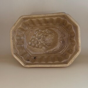 May include: A tan-colored, octagonal ceramic mold with a detailed fruit design. The mold features a central grape and fruit motif, surrounded by a decorative border. The overall shape is rectangular, with a slightly raised design.