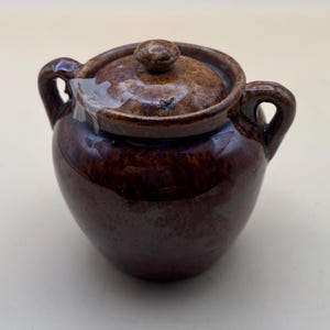 May include: A small brown ceramic pot with a lid and two handles. The pot has a glossy finish and a rustic look.