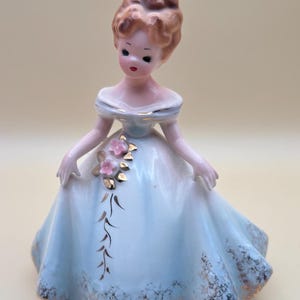 Könnte beinhalten: Eine Porzellanfigur eines Mädchens in einem hellblauen Kleid mit goldenen Verzierungen und rosa Blumen. Das Mädchen hat braunes Haar und trägt einen weißen Kragen.