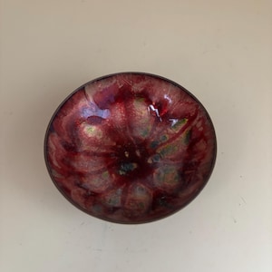 Könnte beinhalten: Eine kleine, runde Schale mit einer rot-gold-grünen Marmorierung. Die Schale ist aus Metall und hat eine glänzende Oberfläche.