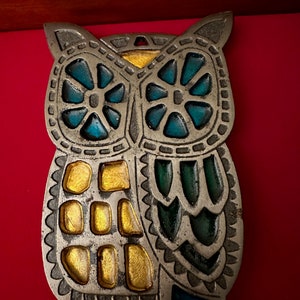 May include: A silver metal owl ornament with blue, green, and gold accents. The owl's eyes are blue, the body is green and gold, and the wings are green.