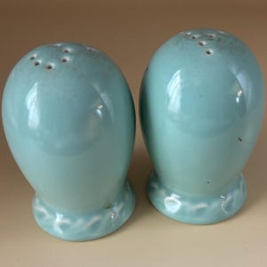 May include: Two matching, light blue ceramic salt and pepper shakers. Each shaker has a rounded, egg-like shape with a series of holes at the top. The base of each shaker has a decorative, wave-like design.