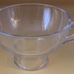 May include: A clear glass teacup with a handle. The cup has a rounded bowl shape and sits on a small, clear base. The handle is a loop shape. The cup is empty and appears to be made of a thick glass material.
