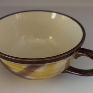 May include: A large ceramic teacup with a wide, open bowl and a brown handle. The interior is cream-colored, while the exterior features a yellow and brown diagonal pattern. The rim and handle are a dark brown.