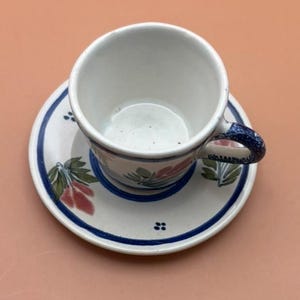 May include: White ceramic teacup and saucer set. The cup and saucer feature a blue border and floral designs in pink, green, and blue. The handle of the cup is blue. The set is on a peach-colored surface.