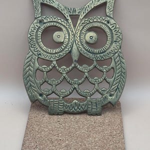 May include: A decorative metal owl-shaped trivet in a bronze finish, with intricate detailing. It sits atop a square cork coaster with a black border. The owl design features large, detailed eyes and a textured surface, suitable for kitchen or home decor.
