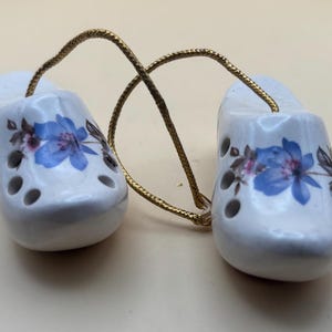 May include: Two white ceramic clogs with blue floral designs and gold string hanging from the top.