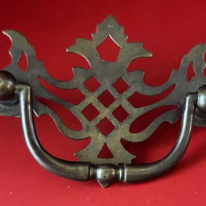 May include: Antique brass drawer pull with an ornate, openwork design. The handle is a curved bar with decorative end pieces. The central design features a diamond pattern and a stylized floral element. The hardware is set against a red background.