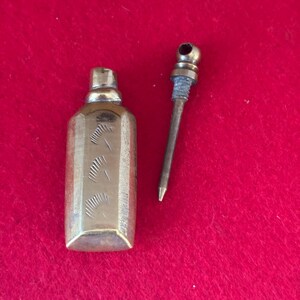 May include: A small, antique brass perfume bottle with a matching applicator. The bottle is rectangular with etched vertical lines. The applicator has a long, thin metal rod with a rounded top. The items are set against a red background.