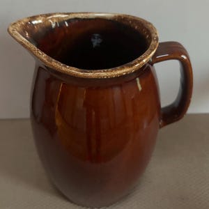May include: A brown ceramic pitcher with a glossy finish. The pitcher has a rounded body, a curved handle, and a spout with a lighter brown rim. The interior is dark. This vintage pitcher is suitable for serving beverages.