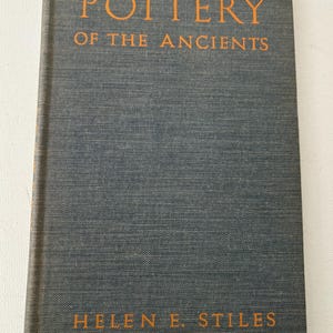 May include: A hardcover book titled "Pottery of the Ancients" by Helen E. Stiles. The book has a dark gray cover with orange lettering. The book's cover has a textured appearance.