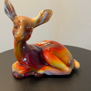 May include: A glass figurine of a deer in a sitting position. The deer is made of red, orange, and yellow glass with white swirls. The figurine is on a black surface.