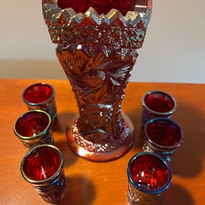 May include: A set of seven red iridescent glass shot glasses and a matching vase. The glasses and vase have a textured, diamond-shaped pattern.