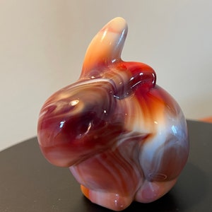 May include: A swirled glass rabbit figurine in shades of red, white, and purple. The rabbit has a long, pointed ear and a rounded body.