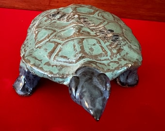 May include: A ceramic turtle figurine with a blue-green glaze and a textured shell. The turtle is standing on a red surface.